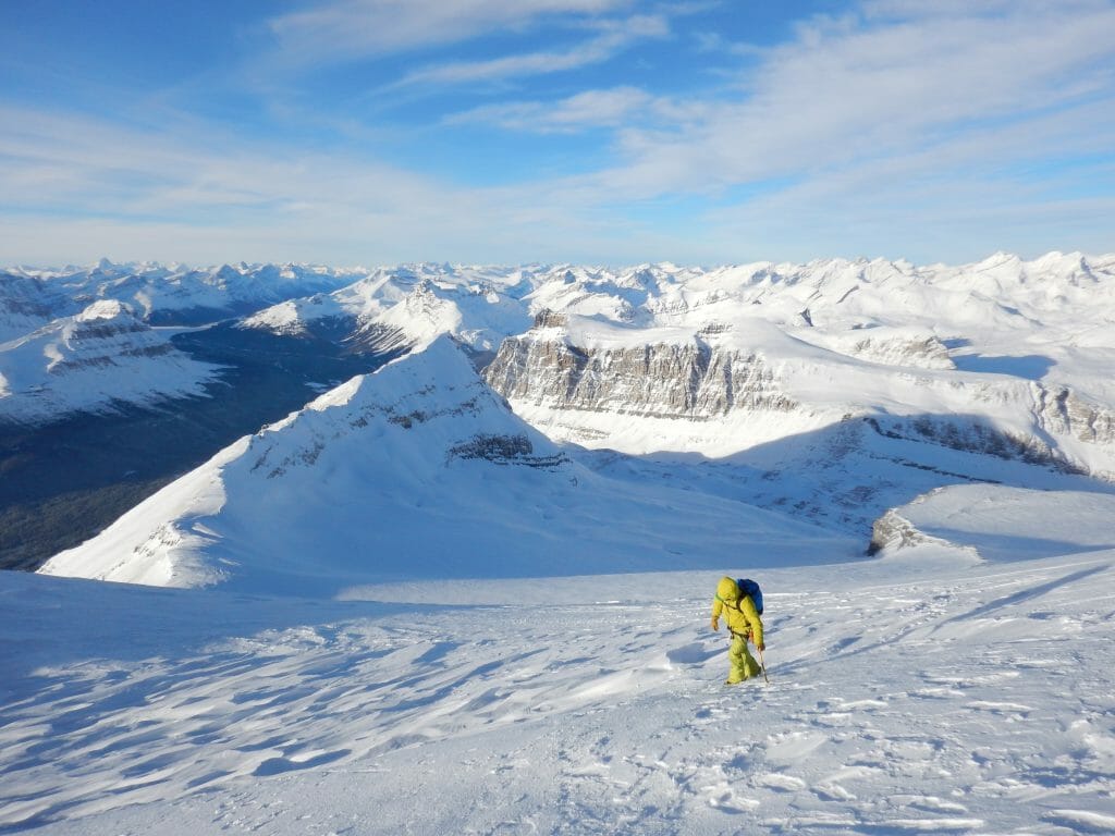A photograph of a man clothed in yellow walking up a steep snow slope.