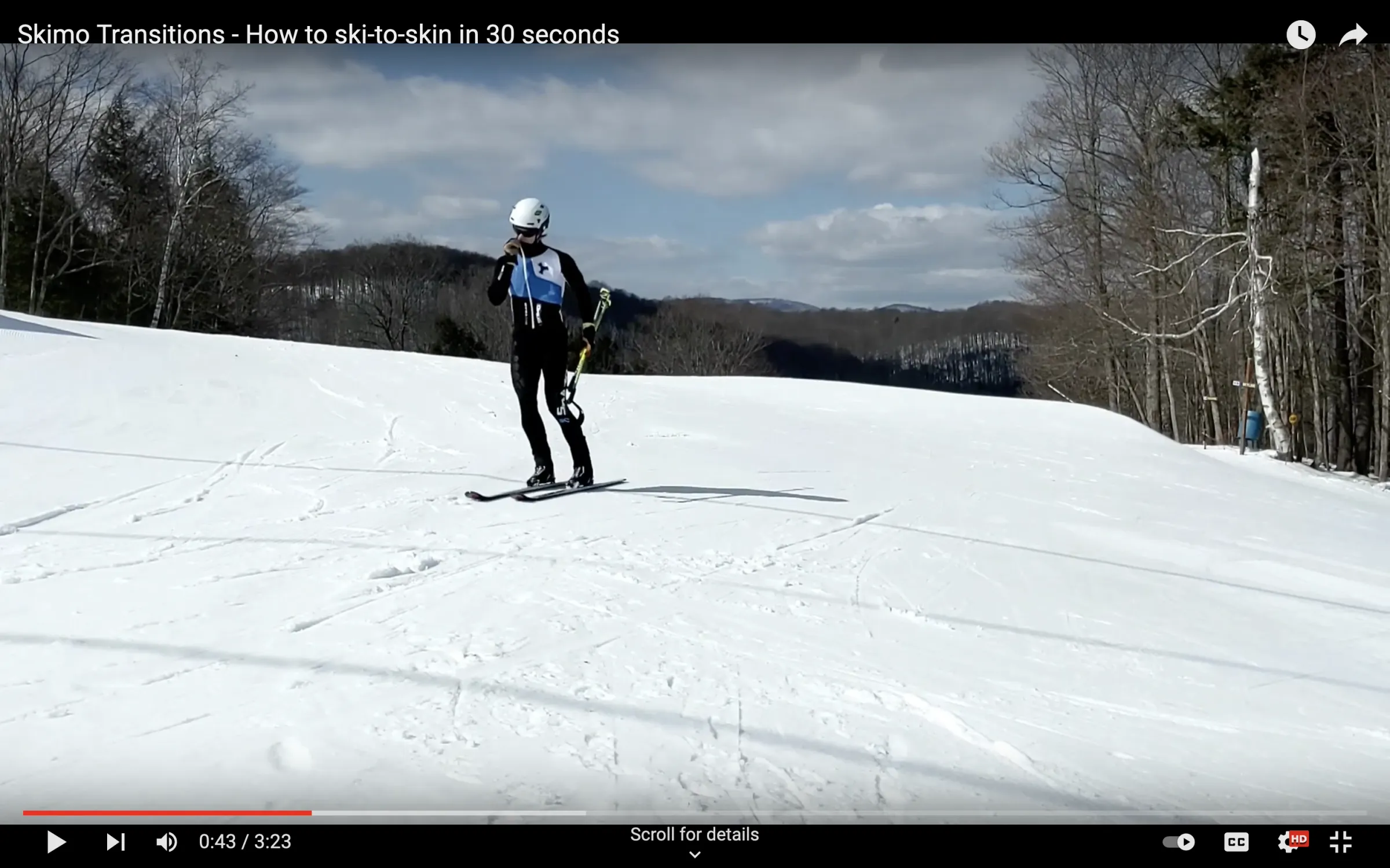 A photo of a skimo racer approaching a transition zone, poles in one hand, other hand on zipper